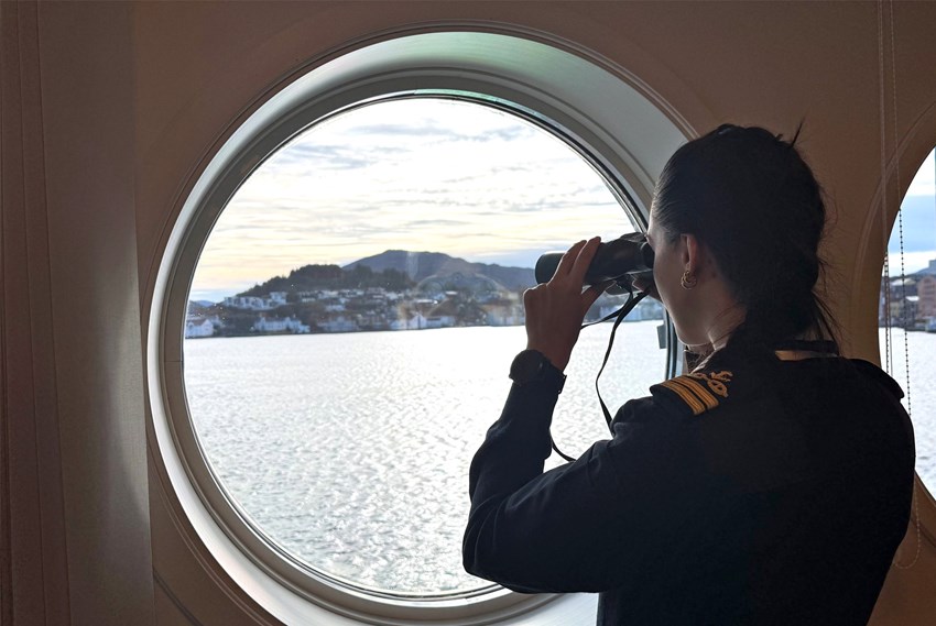Kvinne i uniform ser ut på havna med kikkert. Foto.